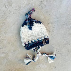 May include: White knitted hat with a blue and purple patterned brim and a long, knotted, blue and purple cord. The hat has a small brown tag with the word "heeb" on it.