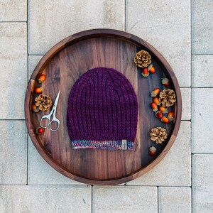 Könnte beinhalten: Eine tiefviolette Strickmütze mit Rippenmuster, die auf einem Holztablett liegt. Die Mütze hat einen bunten gestreiften Saum und ein kleines Etikett. Das Tablett ist mit Tannenzapfen, Eicheln und einer kleinen Schere dekoriert.