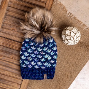 May include: A blue knitted hat with a white and blue floral pattern and a brown faux fur pom pom. The hat has a small brown tag with the word "Heb" on it.