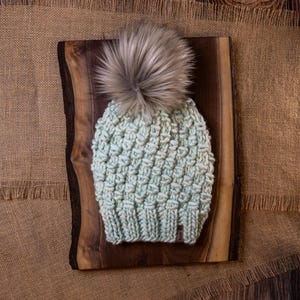 May include: A light blue knitted beanie with a textured pattern and a large gray faux fur pom-pom. The hat is displayed on a wooden board with a natural edge, set against a burlap background.