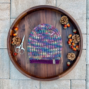Könnte beinhalten: Handgestrickte Beanie-Mütze in leuchtenden Farben, darunter Lila, Blau, Grün und Gelb. Die Mütze hat einen tiefvioletten Rand und ein kleines Etikett. Sie liegt auf einem Holztablett mit dekorativen Tannenzapfen, Eicheln und einer Schere.