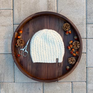 Könnte beinhalten: Hellblau gestrickte Beanie-Mütze mit kleinem braunen Etikett, präsentiert auf einem dunklen Holztablett. Das Tablett ist mit Tannenzapfen, Eicheln und einer kleinen silbernen Schere dekoriert. Der Hintergrund ist eine graue Steinoberfläche.