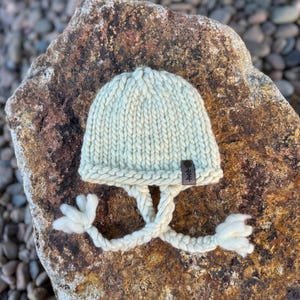 Könnte beinhalten: Eine weiße Strickmütze mit einem langen, geflochtenen Bommel. Die Mütze hat ein kleines braunes Leder-Etikett mit dem Wort "Hee" darauf.