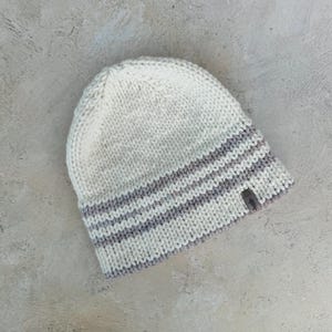 May include: A white knitted beanie with a grey striped band. The beanie has a small brown tag with the word "Happy" on it.