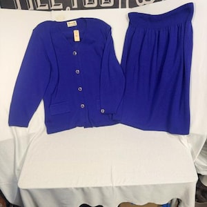 May include: A royal blue two-piece set consisting of a long-sleeved V-neck cardigan with four front buttons and two flap pockets, and a matching high-waisted skirt with an elasticated waistband. The cardigan features a small brand tag at the neck.