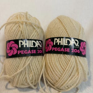Könnte beinhalten: Zwei Knäuel cremeweißes Garn, jeweils mit einer schwarzen Papierbanderole versehen, die das rosa Phildar-Logo und den Text Phildar International Pegase 206 zeigt. Das Garn ist oval gewickelt, mit losen Fäden an der Unterseite jedes Knäuels.