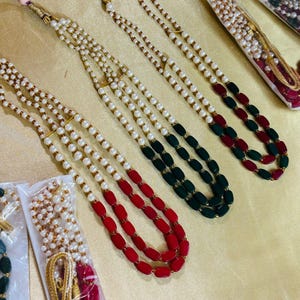 May include: Three strands of pearl necklaces with red and green beads. The necklaces are displayed on a gold background.
