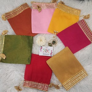 May include: Assortment of seven small fabric pouches with gold trim and tassels. The pouches are in various colors, including red, pink, green, yellow, and orange. The pouches are arranged on a white fluffy surface. A small card with the text "Aishwarya" is visible in the center of the image.