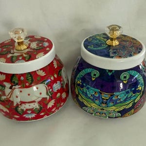 May include: Two decorative ceramic jars with lids. One jar is red with a white and red floral and animal pattern. The other jar is dark blue with a peacock design. Both jars have white rims and clear crystal knobs.