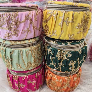 May include: A stack of colorful, round fabric pouches with gold embroidered floral designs. The pouches are in shades of pink, yellow, green, dark green, and orange. Each pouch has a zipper closure and a pleated fabric design.