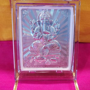 May include: A silver-colored framed image of the Hindu deity Ganesha. The image is set against a white background with a silver-colored sunburst design. The frame is clear plastic.