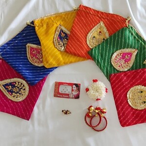 May include: A collection of colorful, patterned drawstring pouches in red, blue, yellow, orange, and green, each with a decorative gold and jewel-toned appliqué. Also included are a box of bindi, floral hair accessories, and red bangles.