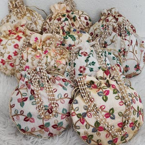 May include: A group of eight small, cream-colored drawstring bags with floral embroidery and pearl embellishments. The bags are arranged in a circle on a white surface.