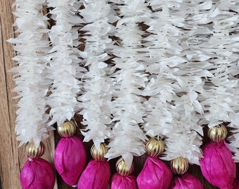 May include: White paper flower garland with pink fabric balls hanging from a wooden wall. The garland is made of multiple strands of paper flowers, each with a gold bead at the bottom.