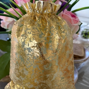 May include: A gold drawstring gift bag with a floral pattern. The bag is made of a shiny fabric and has a gathered top. Pink and white flowers are in the background.