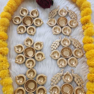 May include: A collection of six gold-colored metal diya oil lamp holders arranged in two columns. Three holders feature a circular flower-like design, while the other three incorporate peacock motifs. The items are displayed on a white surface.