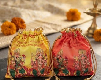 Puede incluir: Dos bolsas con cordón, una amarilla y otra naranja, con ilustraciones impresas de las deidades hindúes Ganesha y Lakshmi. Cada bolsa tiene cierres de cordón dorados con pequeños cascabeles y texto en hindi impreso en la tela.