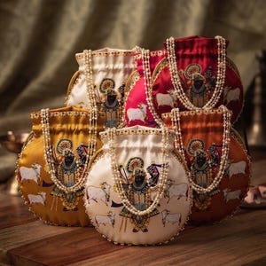 May include: A collection of five round drawstring pouches in cream, gold, red, and orange satin fabric. Each pouch features a printed illustration of a deity surrounded by cows and is adorned with a long, draped strand of white faux pearls.