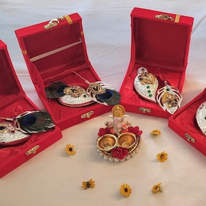 May include: Four red velvet boxes, each open to reveal ornate silver and gold decorative items. Some boxes contain peacock feathers. A central decorative piece and small yellow flowers complete the display.