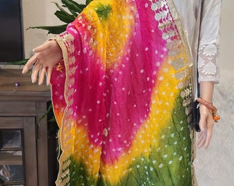 May include: A vibrant, multi-colored scarf with a tie-dye effect. The scarf features a gradient of pink, yellow, and green with white polka dots. The edges are trimmed with gold-colored lace and metallic accents. The scarf is draped over a mannequin.