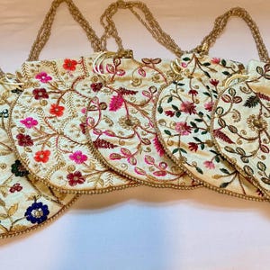 May include: A collection of embroidered drawstring bags in various floral designs. The bags are cream-colored with gold accents and feature colorful floral embroidery. Each bag has a gold chain strap.