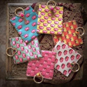May include: A collection of six square fabric pouches featuring a repeating pink lotus flower pattern. Each pouch includes a circular gold-toned metal handle with a small colorful tassel accent. The pouches are available in blue, yellow, orange, light green, pink, and white.