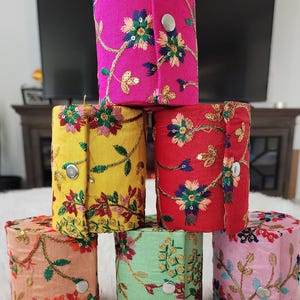 May include: A stack of six cylindrical fabric boxes in various colors, including pink, yellow, red, peach, green, and light pink. Each box is adorned with embroidered floral patterns in complementary colors and secured with a silver snap closure. The boxes are stacked on a white, fluffy surface.