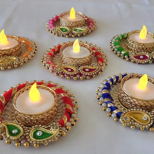 May include: Six decorative candle holders with LED candles. Each holder has a gold-colored metal base with colorful beaded and embroidered accents. The holders are in shades of red, orange, green, and blue, with gold and white accents.