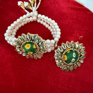 May include: A white pearl bracelet and matching ring set. The bracelet has three strands of pearls and a central decorative element with a green and yellow fabric design, surrounded by clear stones. The ring mirrors the design.