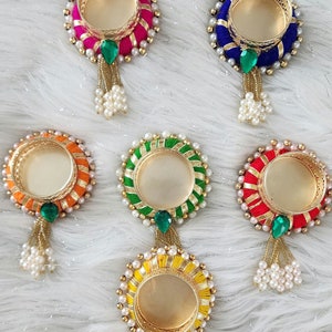 May include: Five round candle holders with gold metal rims and colorful fabric bases. Each holder has a green gem and a pearl tassel.