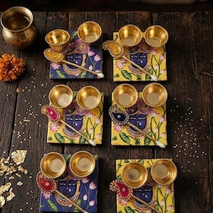 May include: Six square coasters featuring a blue-faced deity illustration with lotus flowers, each paired with two small gold-toned metal bowls and a decorative paisley-shaped spoon with a white tip, arranged in a grid on a dark wooden surface.