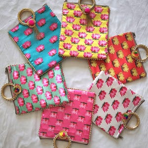 May include: A collection of colorful, patterned clutch bags. Each bag features a lotus flower design in shades of pink and yellow on various backgrounds, including turquoise, yellow, and red. They have gold handles and decorative tassels.