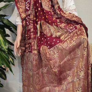 May include: A maroon and gold patterned textile, possibly a shawl or scarf, draped over a mannequin. The fabric features intricate gold floral and paisley designs on a deep red background. The edges are trimmed with a gold border.