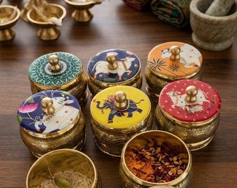 May include: A collection of round brass containers with decorative lids featuring colorful painted illustrations of cows, elephants, and floral mandalas. Each container has a small central knob handle and contains various spices and grains.