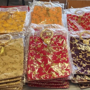 Puede incluir: Una colecci&oacute;n de peque&ntilde;as bolsas decorativas con bordados dorados y borlas. Las bolsas est&aacute;n hechas de una tela suave y texturizada y tienen un asa de anillo. Las bolsas est&aacute;n dispuestas en filas sobre una mesa.
