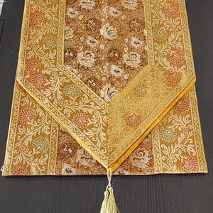 May include: A decorative table runner in shades of gold, brown, and cream. The runner features a floral pattern in the center and ornate borders. A folded section with a tassel adds a decorative element. The runner is designed for home decor.