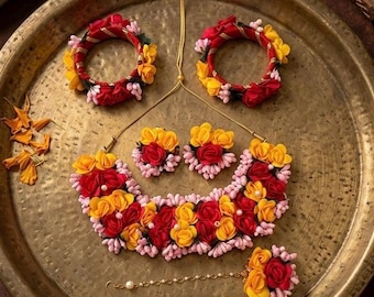 Puede incluir: Un conjunto de joyería que incluye dos brazaletes, un collar, un par de pendientes y un maang tikka, adornados con flores artificiales rojas, amarillas y rosa claro. Las piezas están dispuestas sobre una bandeja de metal martillado.
