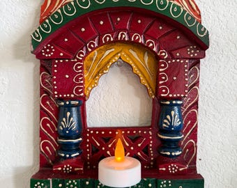 Puede incluir: Un marco decorativo de madera para pared con diseño de ventana arqueada y detalles pintados a mano en rojo, verde, azul y amarillo. La estructura incluye dos pilares azules, un motivo de cortina amarilla y una pequeña vela LED blanca.