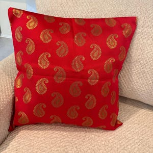 May include: A red decorative pillow with a gold paisley pattern. The pillow is rectangular and has a soft, textured appearance. The paisley design is evenly distributed across the surface, creating a visually appealing aesthetic.