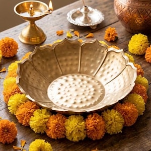 May include: A gold-toned metal bowl with a scalloped edge and a textured, dimpled interior surface. The bowl is surrounded by a circular arrangement of bright yellow and orange marigold flowers, creating a vibrant floral border around the base.