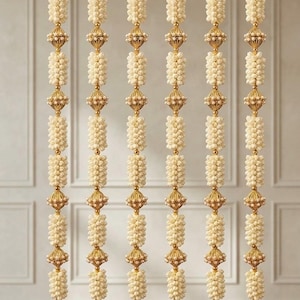 May include: Six vertical hanging garlands featuring alternating segments of cream-colored floral clusters and metallic gold beads. Each strand is topped with a circular gold ring and finished at the bottom with a decorative gold bell-shaped ornament.