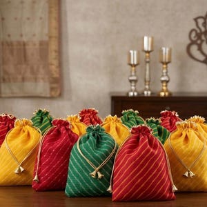 May include: A collection of small drawstring pouches in red, yellow, and green fabric with a diagonal striped pattern. Each pouch features a gold-toned cord closure with two small bell-shaped charms at the ends, arranged on a wooden surface.