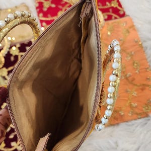 Puede incluir: Un bolso de mano dorado con cierre de cremallera y asa de cuentas. El bolso est&aacute; forrado con un tejido beige.