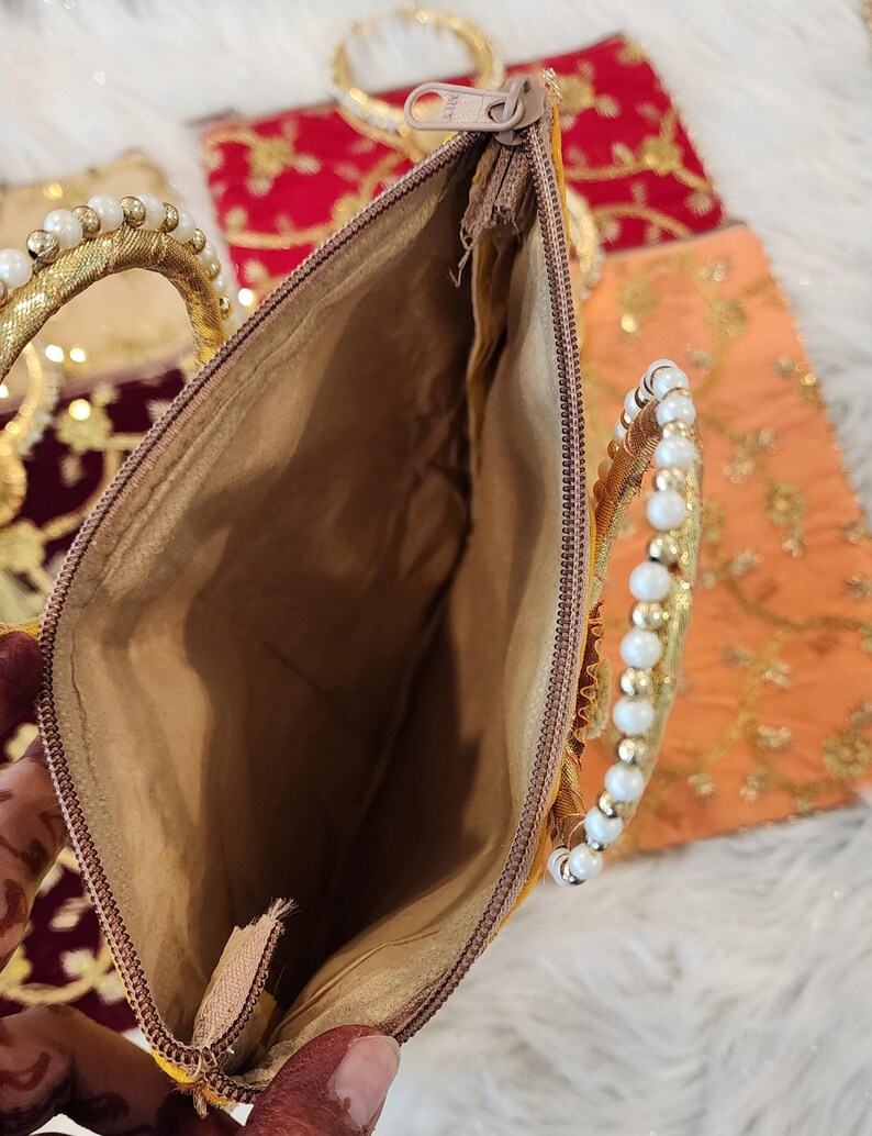 Puede incluir: Un bolso de mano dorado con cierre de cremallera y asa de cuentas. El bolso est&aacute; forrado con un tejido beige.