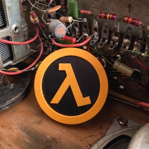 May include: A circular emblem featuring a bright orange Greek lambda symbol centred on a matte black background, surrounded by a thick orange border. The object is positioned in front of an exposed electronic circuit board with various components.