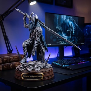 May include: A detailed statue of Artorias the Abysswalker, a character from the Dark Souls video game series. The figure is clad in dark armor and holds a large sword and spear. The base is brown with a gold nameplate. The statue is on a desk with a computer.