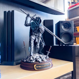 Puede incluir: Una estatua detallada de Artorias el Caminante del Abismo, un personaje de videojuego, sobre una base rocosa con dos espadas. La figura está vestida con armadura plateada y una capa con capucha azul, sosteniendo una espada grande. La base tiene un borde marrón con el nombre del personaje.