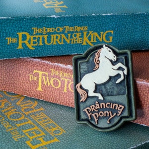 May include: A green, brown, and white enamel pin featuring a white horse with a brown mane and tail. The pin has a green border and the words "Prancing Pony" in white lettering.