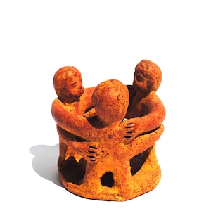May include: A circular sculpture featuring three orange-toned figures standing in a ring, each with arms extended to embrace the others. The textured, speckled surface of the figures creates a uniform appearance across the entire connected group.
