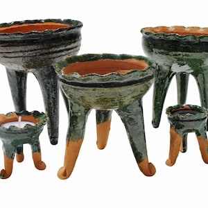 Puede incluir: Una colección de cinco recipientes trípodes de varios tamaños, con un exterior esmaltado en verde oscuro, borde ondulado y una base e interior de terracota sin esmaltar. Cada recipiente se sostiene sobre tres patas de arcilla.